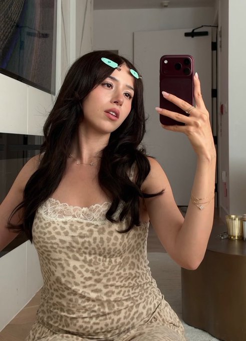 A mirror selfie of a young woman taking a photo with a maroon smartphone; she is wearing a leopard print corset top with lace trim and has a blue hair clip in her dark hair.