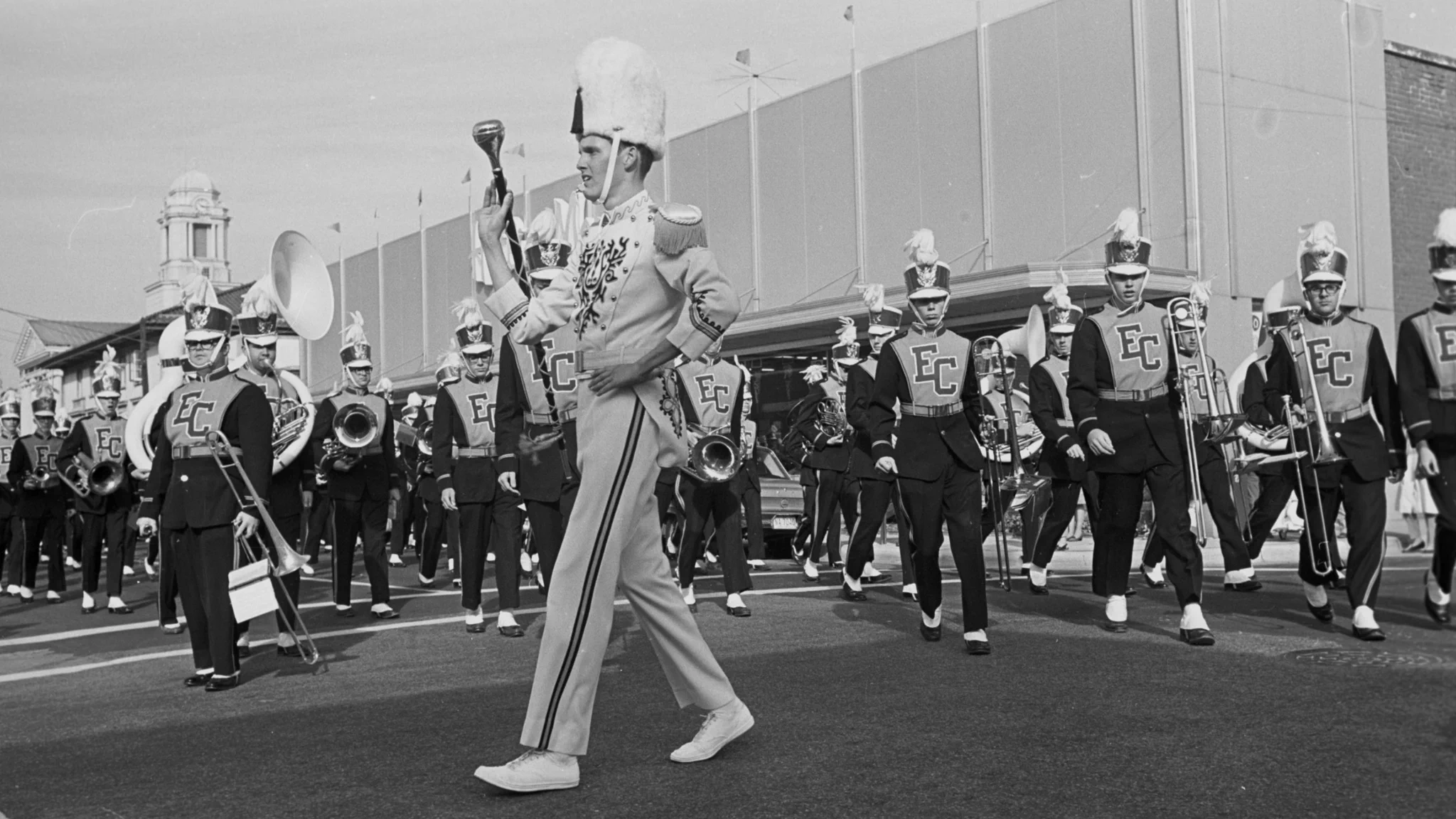 Black-and-white photo of a marching band parading down a street. A drum major in a light uniform with a tall fur hat leads the band. Other band members follow in dark uniforms with \"EC\" emblazoned on their chests, playing brass instruments. Buildings are in the background.