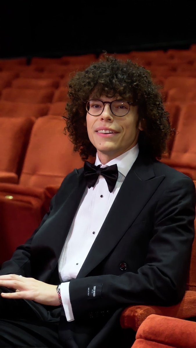 Reece Feldman, the 99th Oscars red carpet correspondent, sits smiling in a formal black tuxedo with bow tie and glasses, posing in a theater with rows of red seats behind him as he prepares to cover the awards ceremony hosted by Conan O'Brien on March 14th.