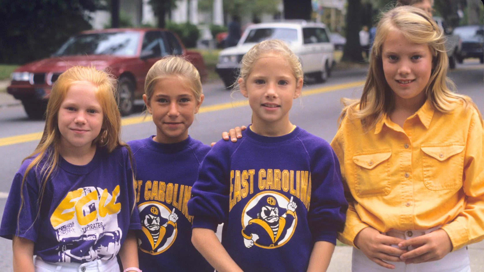 Four smiling girls stand by a street. Three girls wear purple shirts/sweatshirts: one says \"ECU PIRATES\"; two say \"EAST CAROLINA\" with a pirate mascot. The fourth girl wears a yellow shirt.