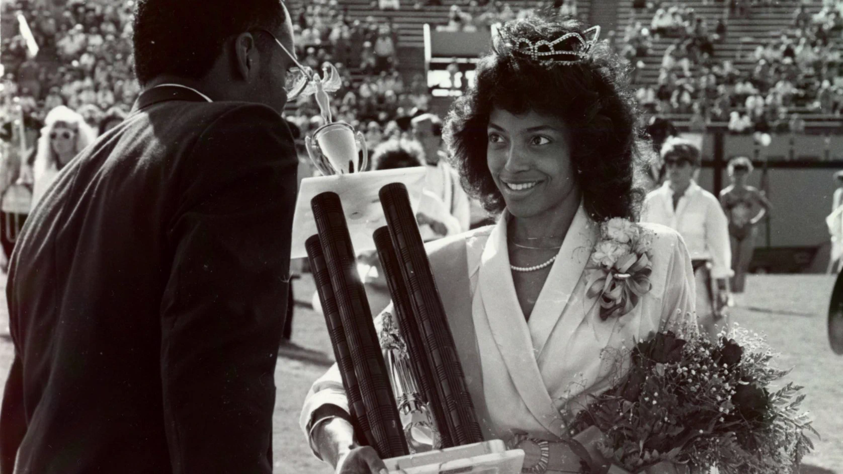 A smiling woman in a crown, pearl necklace, and formal dress holds a bouquet of roses and a winged trophy. She faces a man in a suit, with his back to the camera, wearing glasses. Spectators fill the stadium in the background.