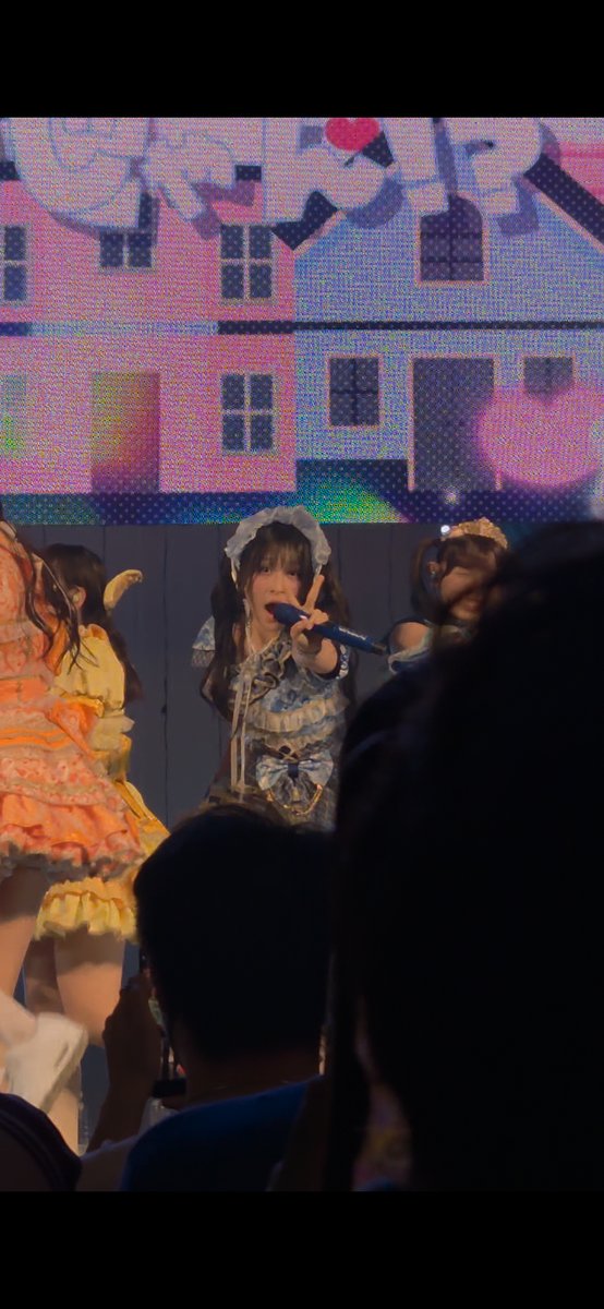 Three performers in elaborate frilly outfits perform on stage in front of a pixelated house backdrop, with one holding a blue microphone and making a peace sign.