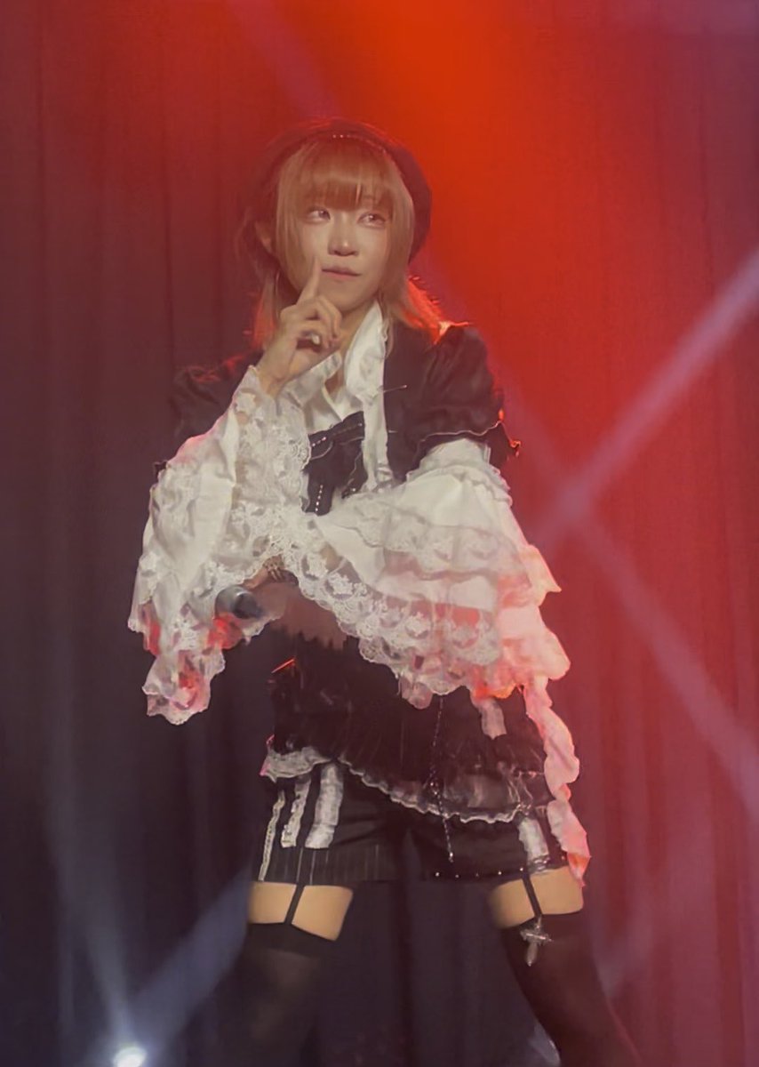 A cosplayer on stage under red lighting wears an elaborate black and white outfit with large ruffled lace sleeves, making a "shh" gesture with one finger raised to their lips.