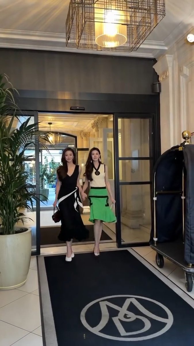 Two women—one in a black dress and the other in a cream top and green skirt—walk out of a building through a doorway onto a black mat displaying a stylized logo.