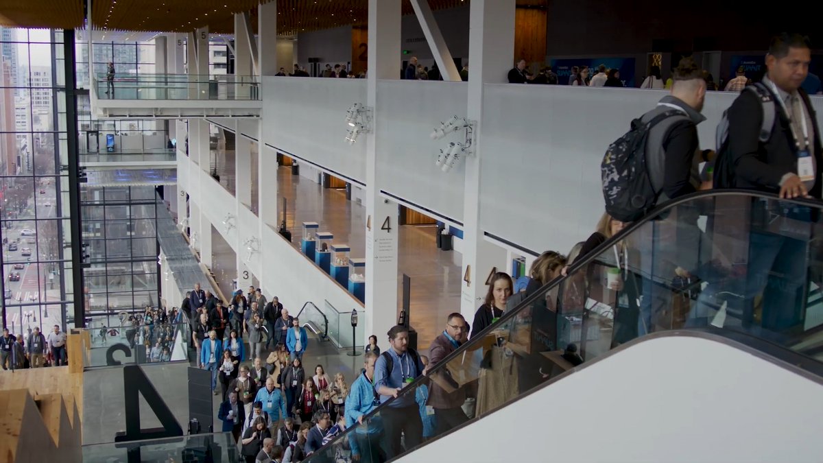 A crowded convention center lobby with people walking and riding escalators, featuring large floor-to-ceiling windows overlooking a city street.