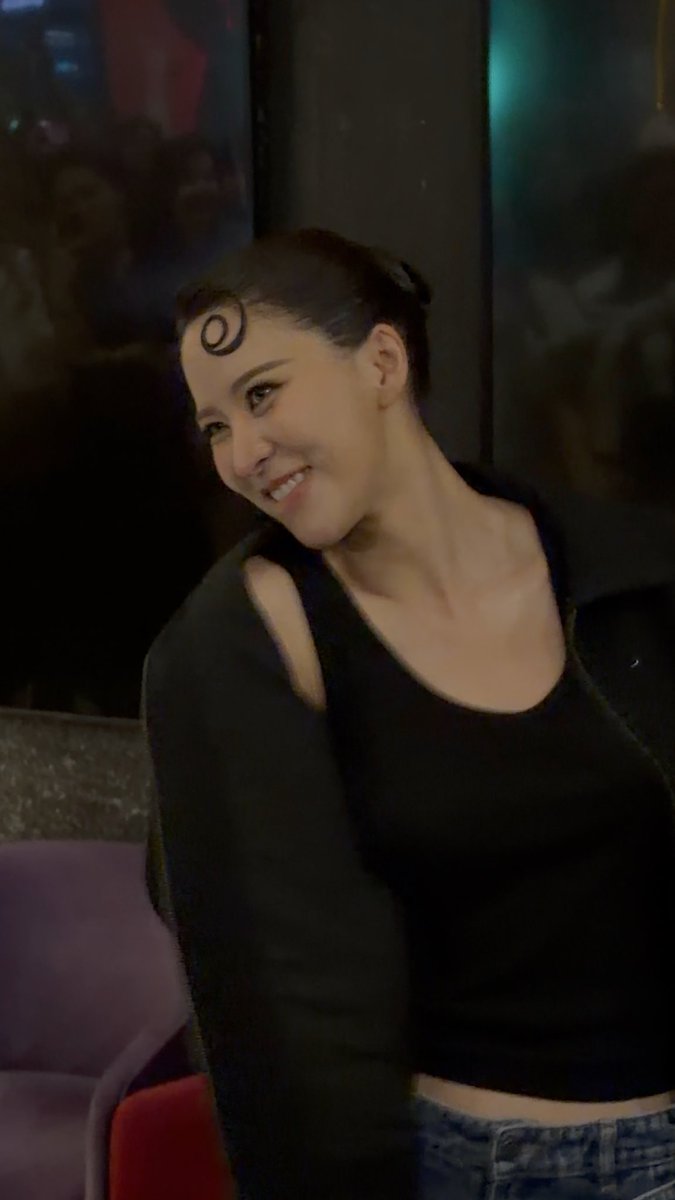A smiling young woman with a crescent moon mark on her forehead and a small cut on her lip poses in a dark environment.