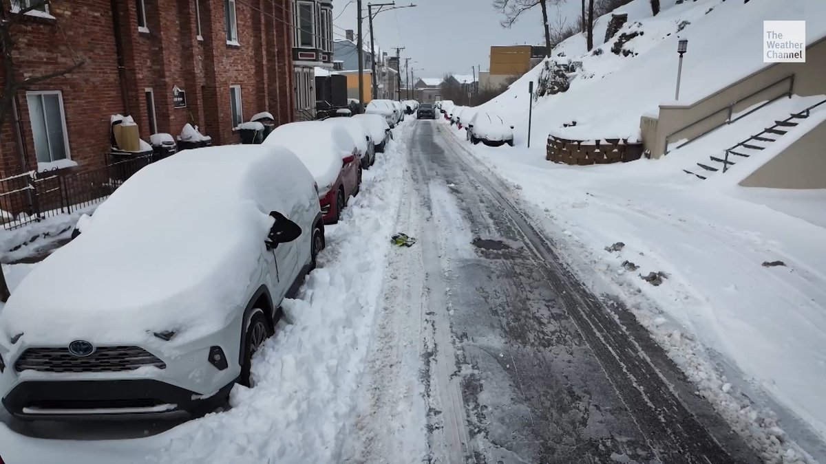 A snow-covered street with a row of parked cars blanketed in heavy snow, including a white Toyota SUV in the foreground, illustrates the lingering aftermath of Winter Storm Fern, which led to half the U.S. declaring states of emergency and prompted the storm's name to be retired from the naming rotation. "The Weather Channel" logo is visible in the upper right corner.