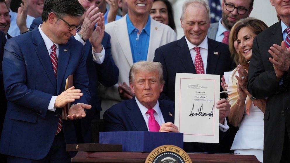 A man seated at a signing desk holds up a signed document while people in formal clothing applaud around him. A presidential seal is visible on the desk, and the document contains text.
