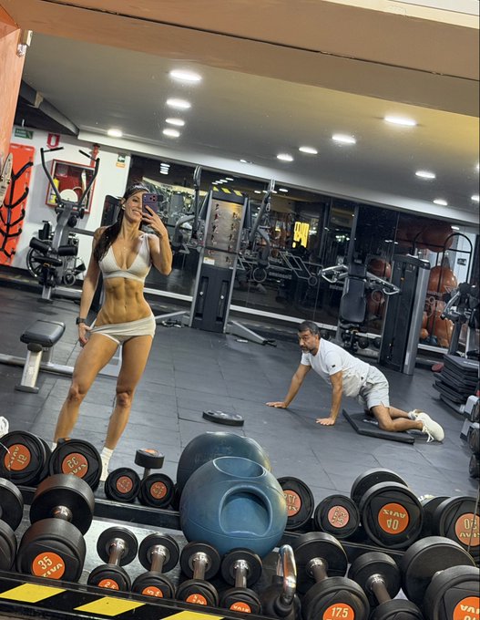 A woman in a white bikini takes a mirror selfie in a gym, displaying her lean physique, while a man does push-ups on a mat in the background, surrounded by dumbbells labeled with weights like "17.5" and "20" and a blue BOSU ball—capturing a humorous moment of contrast that fits the "I don't mind" caption.