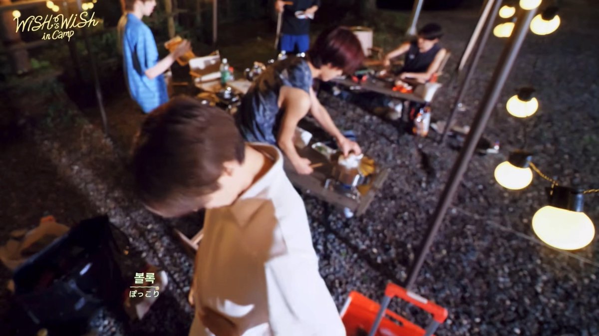 Several young men are gathered around a campsite cooking area at night, with string lights hanging nearby and 'WiSH WiSH in Camp' displayed in the upper left corner.