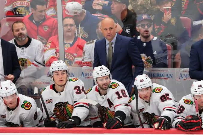 The Chicago Blackhawks bench shows players in white jerseys and a coach in a suit looking on during a hockey game.