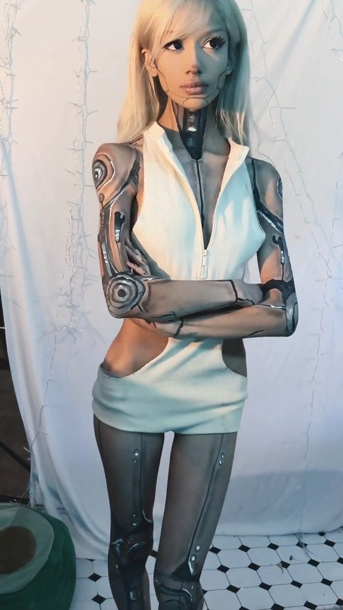 A person cosplaying as a cyborg or robot character with blonde hair, wearing a white sleeveless zip-up dress, posing with arms crossed in a bathroom with tiled flooring.