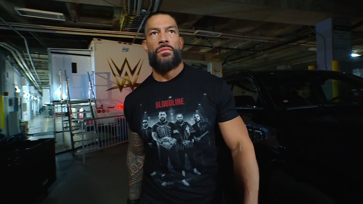 Professional wrestler Roman Reigns stands in a dimly lit parking garage wearing a black 'BLOODLINE' t-shirt, with a WWE production truck visible in the background.