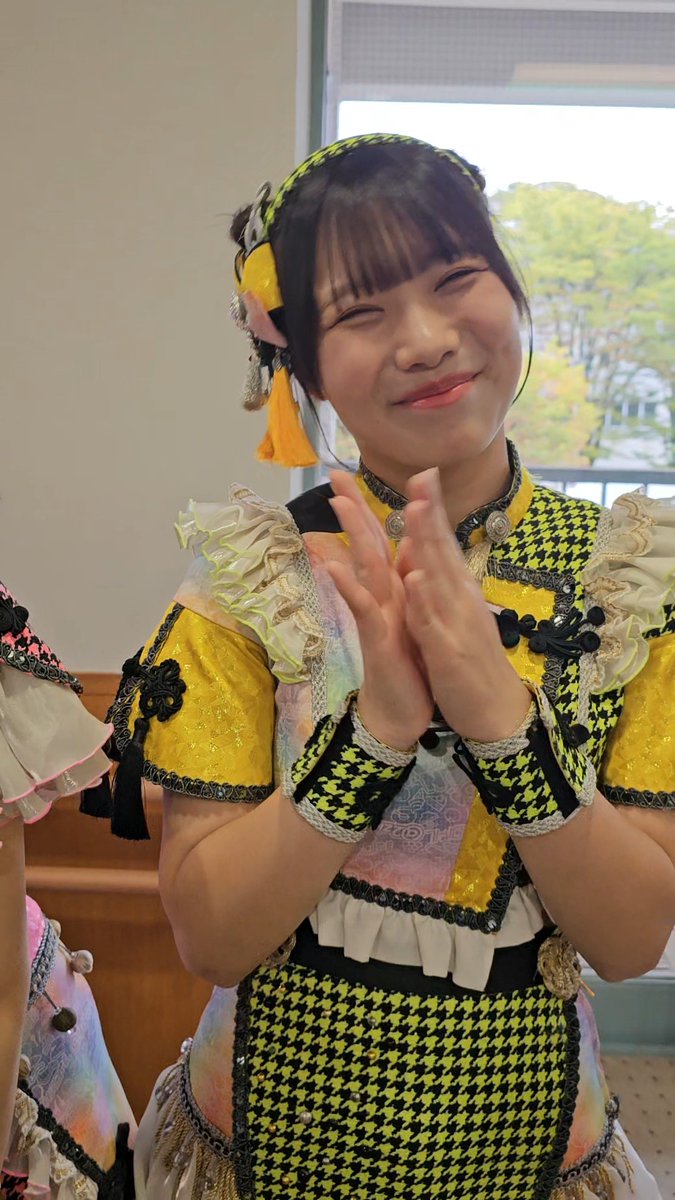 A young woman smiles brightly with her hands clasped together, wearing a vibrant yellow and black houndstooth patterned outfit with ruffles and a matching headband.