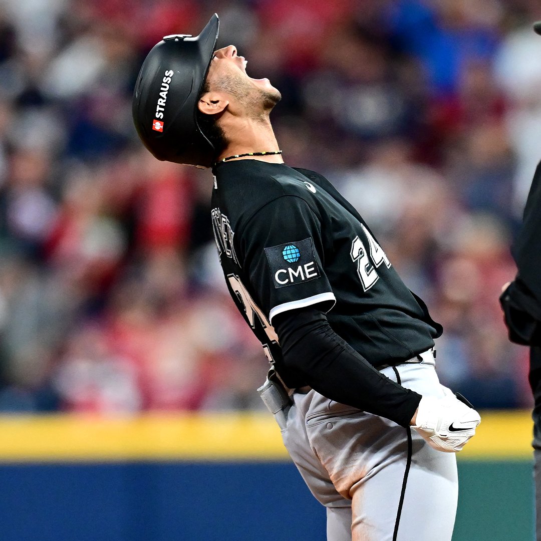 A Chicago White Sox baseball player, wearing a black jersey with a CME patch and a helmet labeled 'STRAUSS,' looks up towards the sky in apparent frustration or celebration.