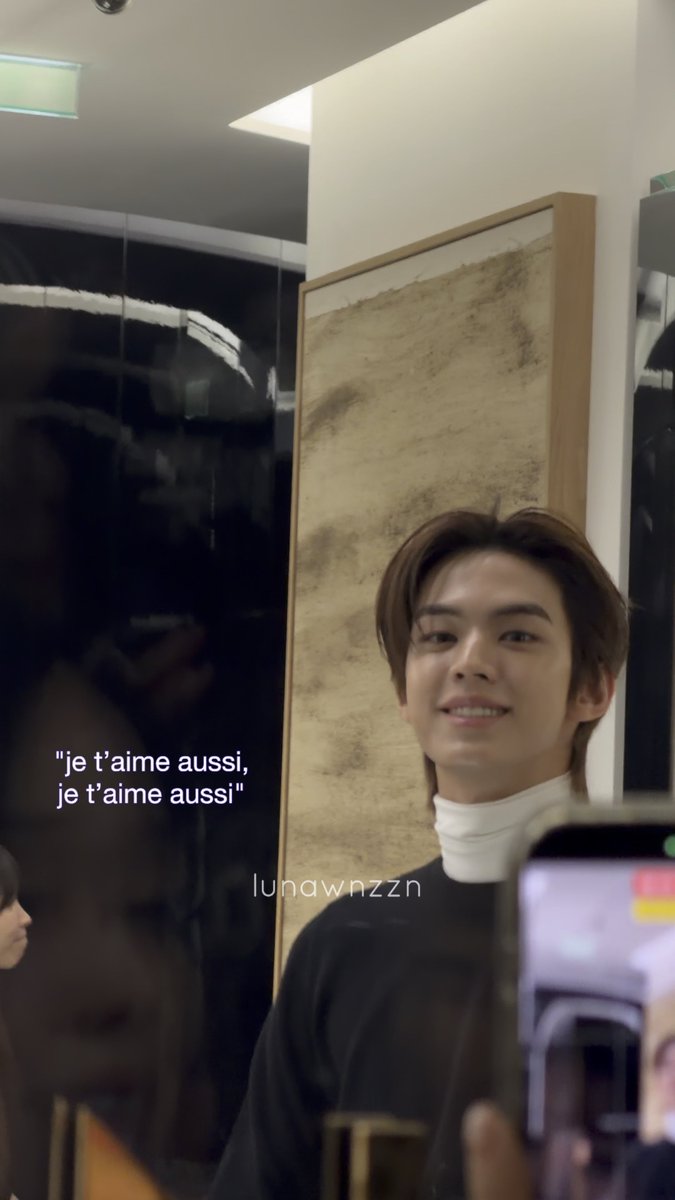 A young man smiling at a camera in a dimly lit room with an abstract painting in the frame, overlaid with the French text 'je t'aime aussi, je t'aime aussi' and the watermark 'lunawnzzn'.