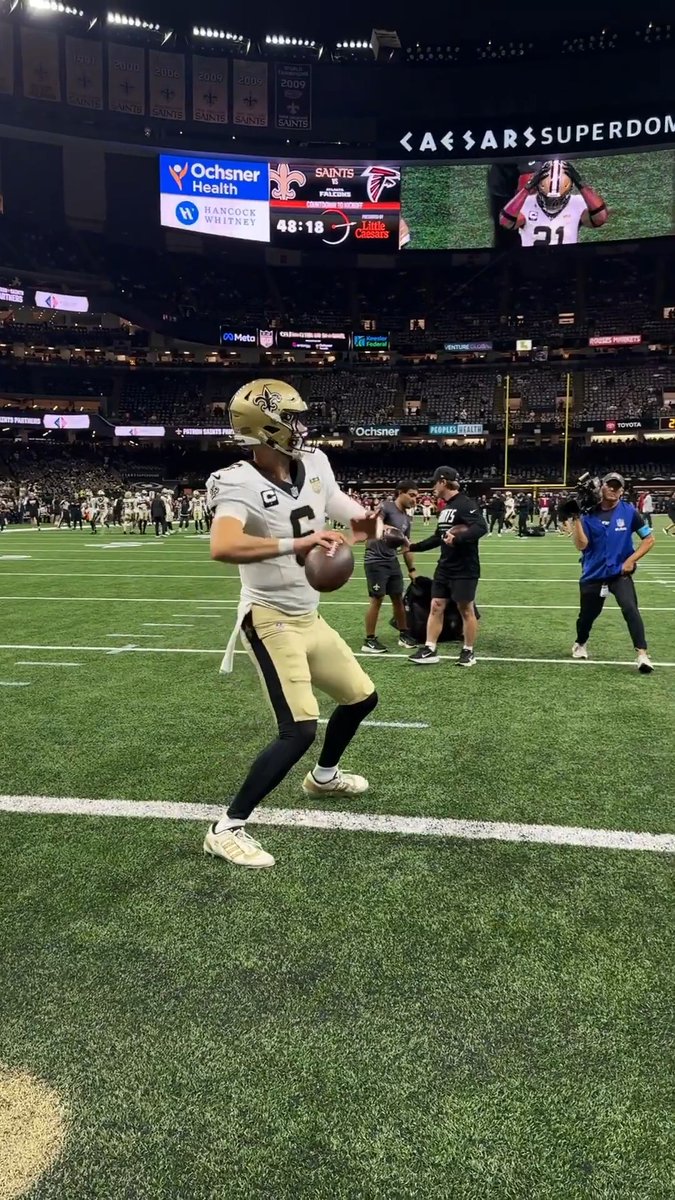 New Orleans Saints quarterback Tyler Shough (wearing jersey #6 with a captain's "C" patch) holds a football on the field at the Caesars Superdome during pregame warmups, with the scoreboard showing the upcoming matchup against the Atlanta Falcons.