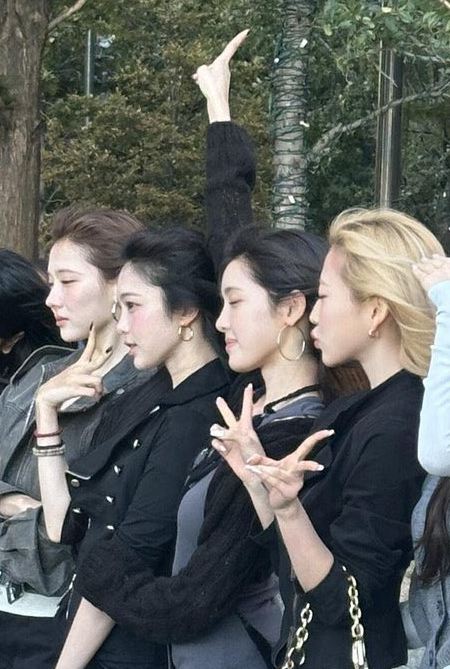A group of young women posing together outdoors, mostly in profile view, with one pointing upward and another making a finger heart gesture, wearing dark and coordinated outfits.