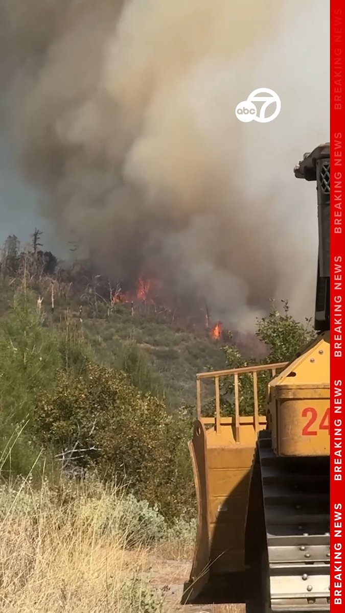 A news broadcast screenshot from ABC7 showing a wildfire burning on a hillside with thick smoke rising into the sky, with a piece of heavy equipment visible in the foreground and 'BREAKING NEWS' banner along the right side.