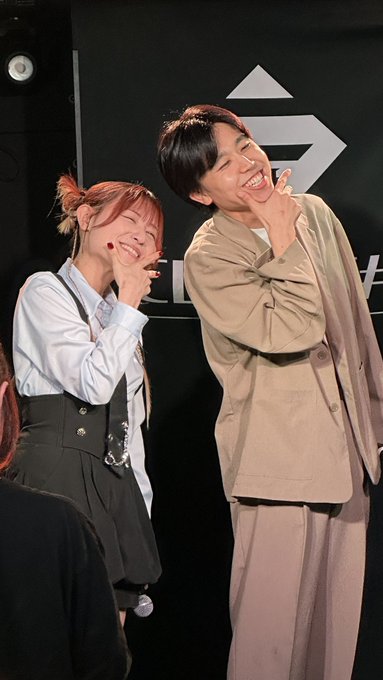A smiling man and woman posing together, both making peace signs with their fingers, appearing to be at an event.