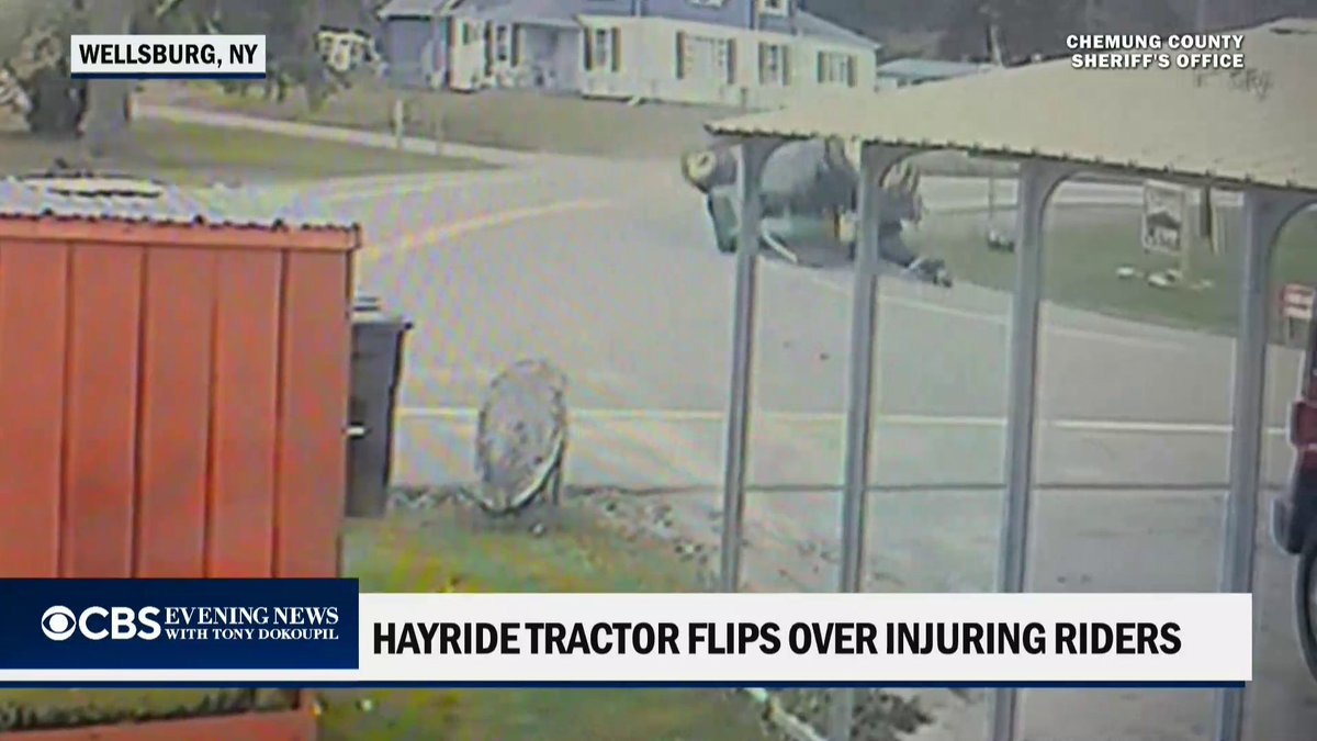 A surveillance video screenshot from the Chemung County Sheriff's Office, labeled "WELLSBURG, NY," shows a tractor mid-rollover on a residential street next to a structure, with the CBS Evening News headline "HAYRIDE TRACTOR FLIPS OVER INJURING RIDERS" displayed below, illustrating the moment the hayride tractor crashed after its brakes failed, injuring the 16-year-old driver and 15 passengers.