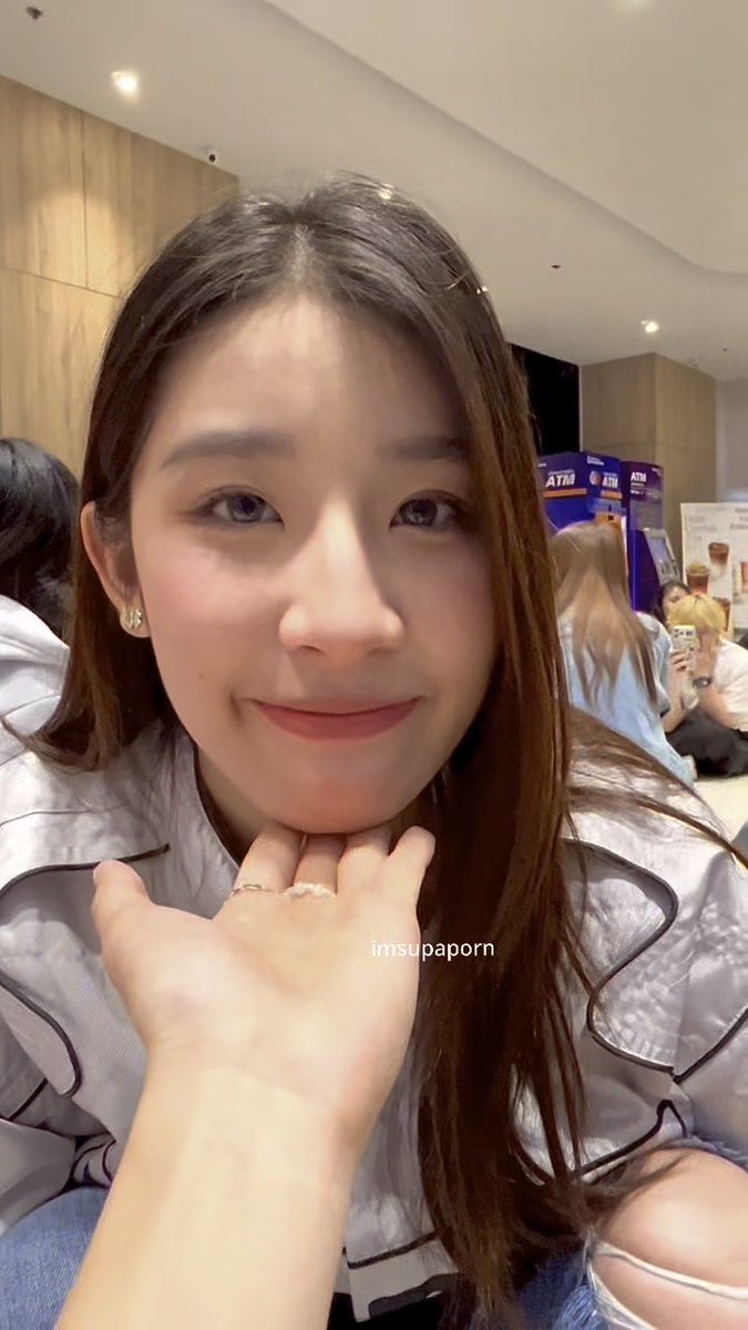 A close-up selfie of a young woman with long brown hair smiling gently in what appears to be a public indoor space with other people in the background, with a watermark reading 'imsupaporn'.