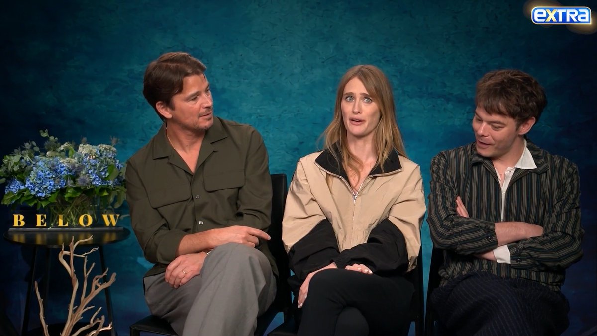 Josh Hartnett, a woman, and Charlie Heaton sit together for an interview promoting their Netflix film "Below," with the title displayed beside a floral arrangement. The "Extra" TV logo appears in the top corner as the actors discuss their Newfoundland accents for the project.