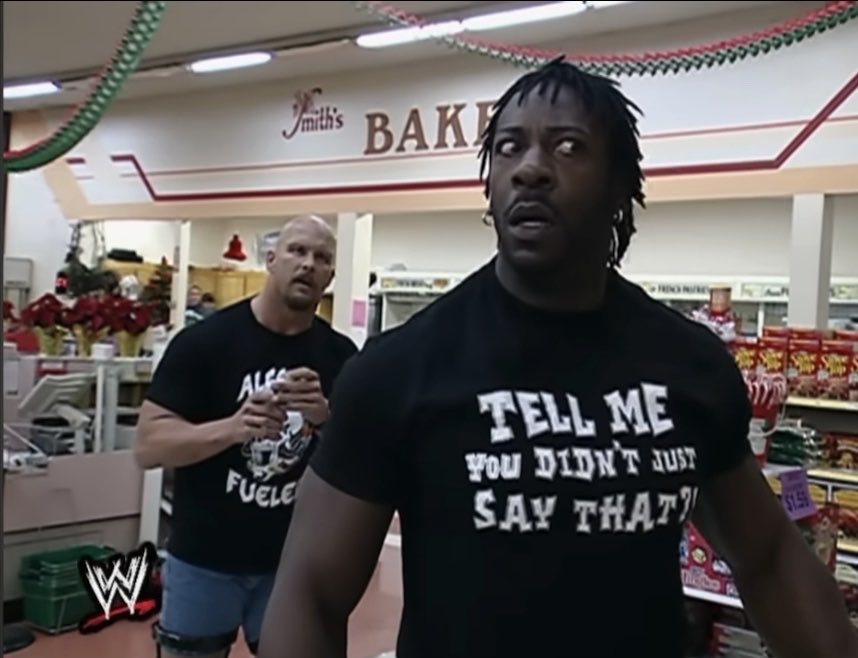 Two WWE wrestlers appear in a Smith's Bakery in a supermarket setting. The man in the foreground wears a black shirt reading 'TELL ME YOU DIDN'T JUST SAY THAT!!' while the muscular bald man in the background wears an 'ALF... FUELED' shirt.
