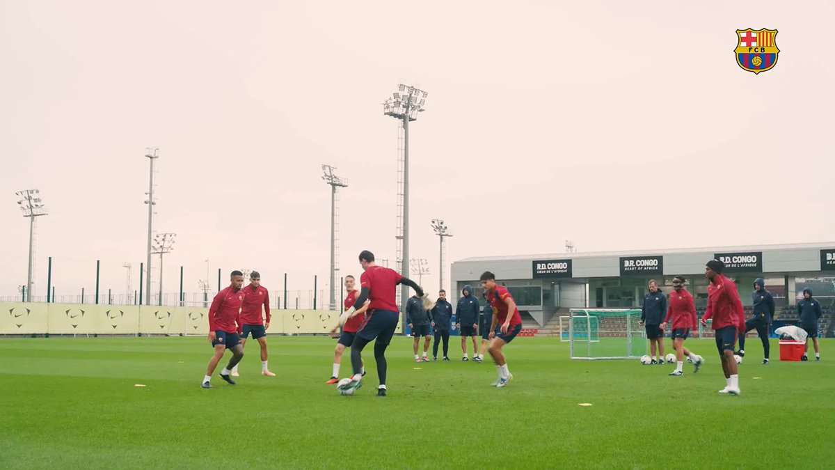 Barcelona players, in red training tops, gather on a green pitch for rondo drills near a facility marked "R.D. CONGO - HEART OF AFRICA," with the FCB crest displayed in the corner. The overcast training ground and small-sided goals complete the team's preparation session.