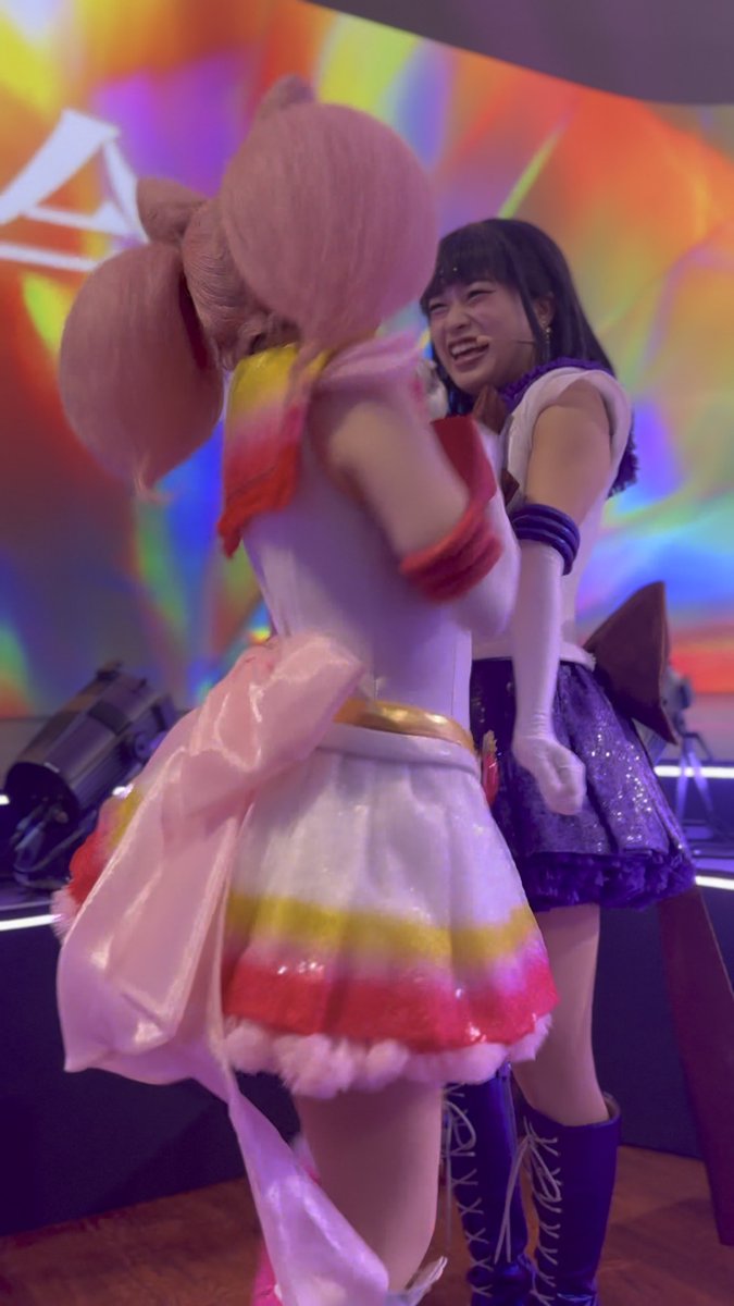 Two performers in Sailor Moon and Chibi Moon costumes interacting on a stage with a colorful rainbow background.