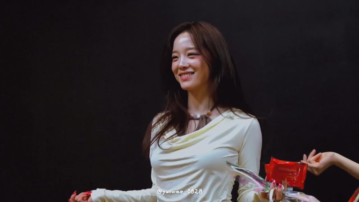 A smiling woman with long dark hair, wearing a white draped top and a beaded necklace, is handed a red package during what appears to be a fan event.