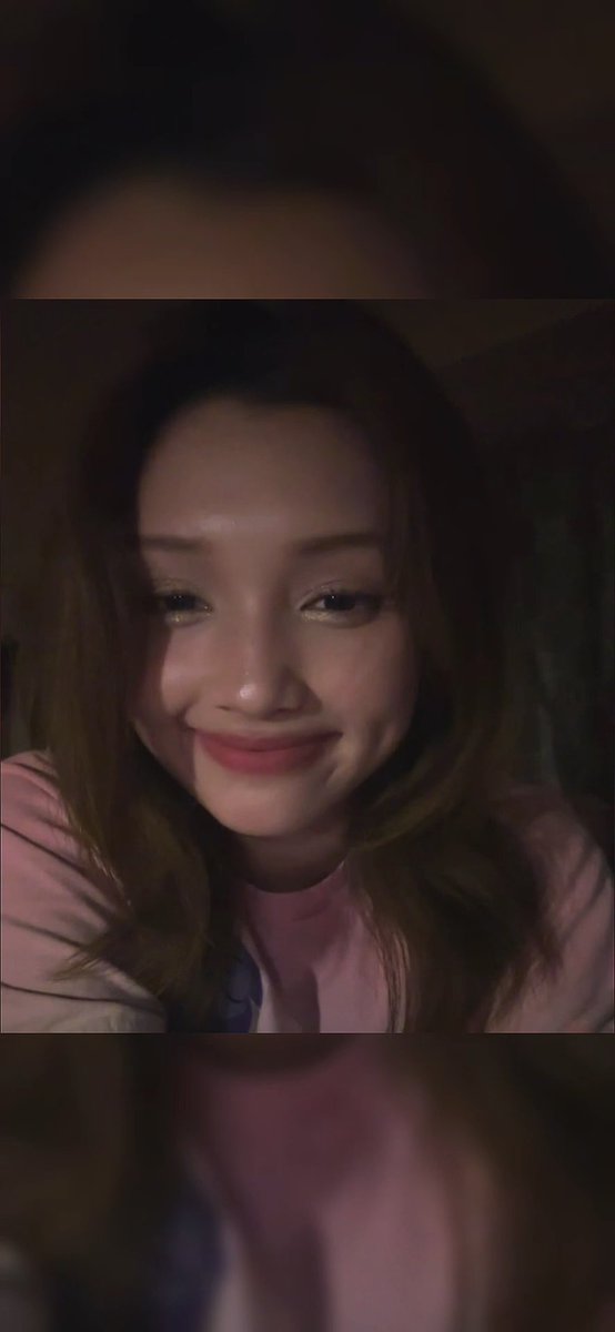 A dark selfie video screenshot of a smiling young woman with long brown hair wearing a pink shirt.