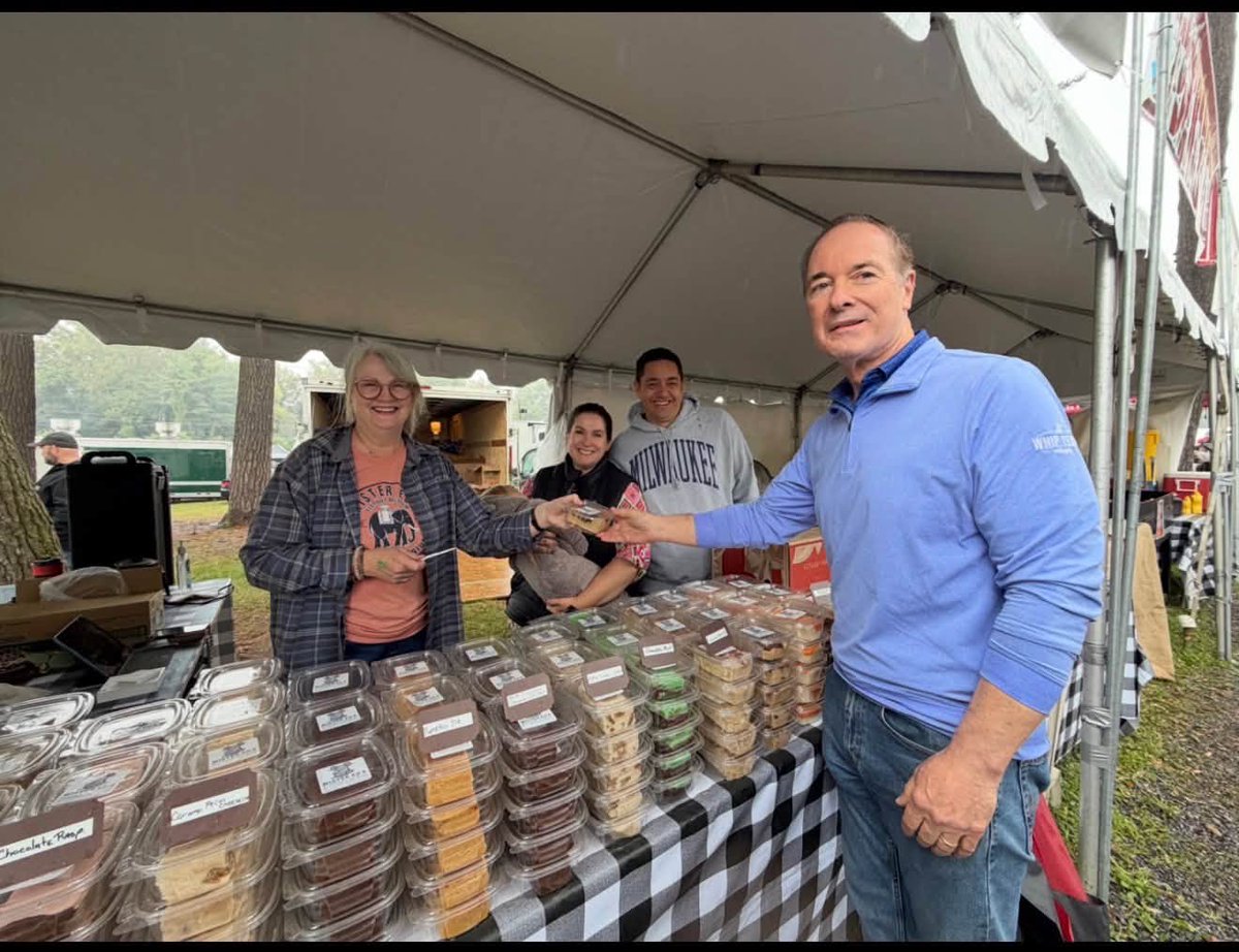 A man in a blue shirt reaches across a market stall to accept or hand over a small item, while three vendors smile behind a table covered with packaged baked goods. The stall is under a tent, and some text appears on clothing and product labels.
