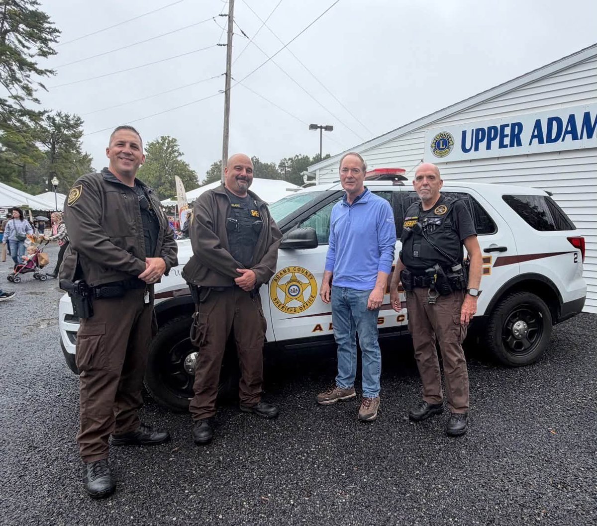 Four men pose for a photograph beside a sheriff's SUV at an outdoor community event. Three men wear sheriff's uniforms, and one wears a blue shirt and jeans. A building with a Lions Club sign, tents, and people are visible in the background. Text appears on the building, uniforms, and vehicle.