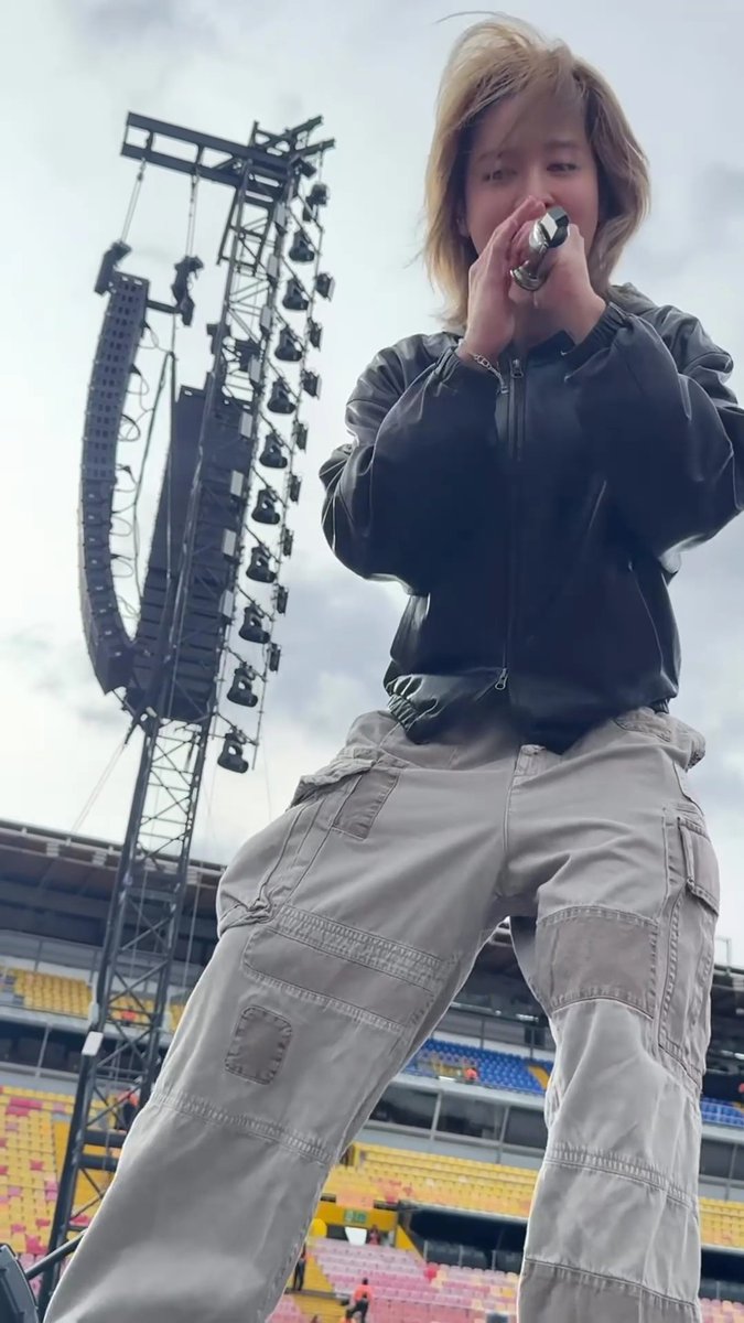 A young blonde-haired person holding a microphone on what appears to be a stadium stage, wearing a black leather jacket and grey cargo pants, with stage lighting and empty colored stadium seats in the background.