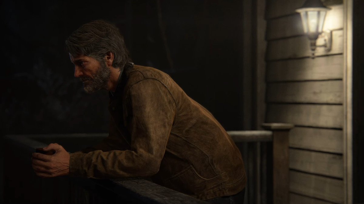 A gruff man with a beard and weathered face in a brown suede jacket leans on a wooden railing at night in front of a house with a lit porch lamp.