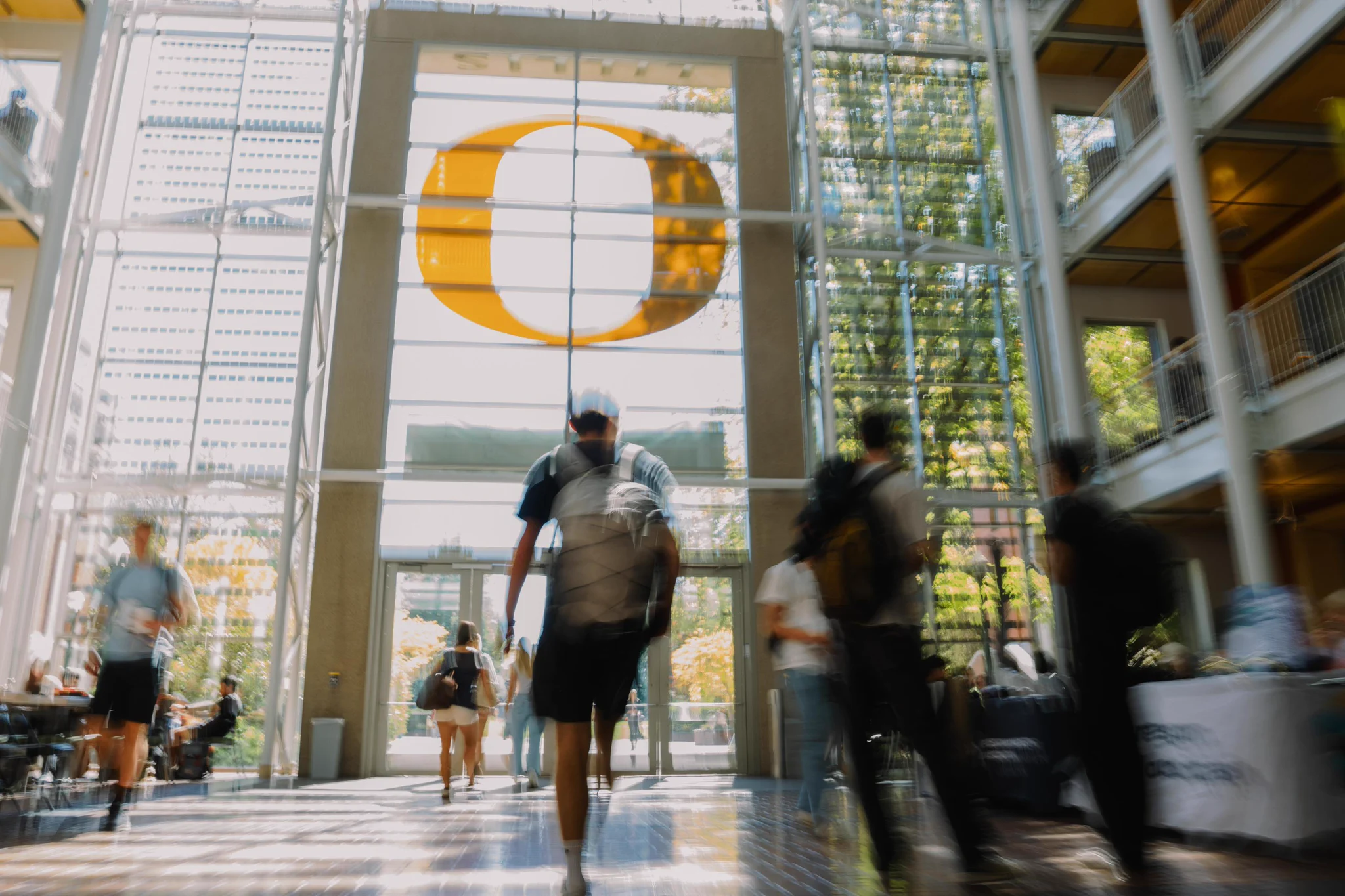 A person walks through a sunlit atrium with a large yellow "O" symbol, associated with the University of Oregon, in the background. Other people are also visible, creating a dynamic, busy scene.
