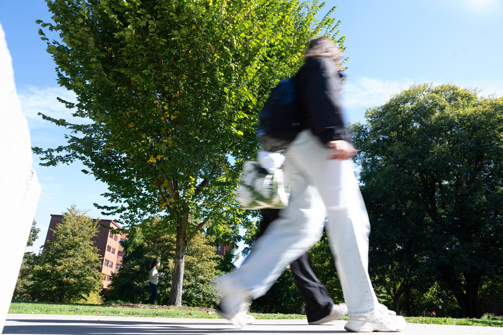 A person walks briskly on a path in a park-like setting with trees and a building visible in the background. The scene is bright and sunny, indicating daytime.