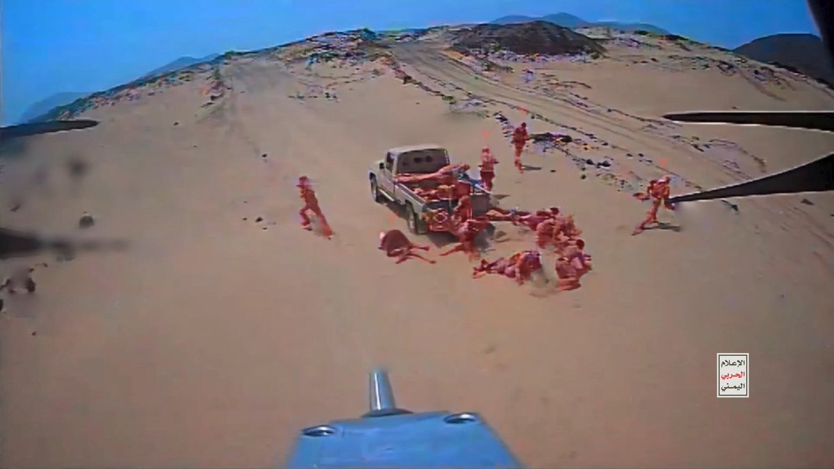 An aerial view shows a group of people gathered around a white pickup truck in a sandy desert landscape, with Arabic text "الإعلام الأمني" visible in the bottom right corner.