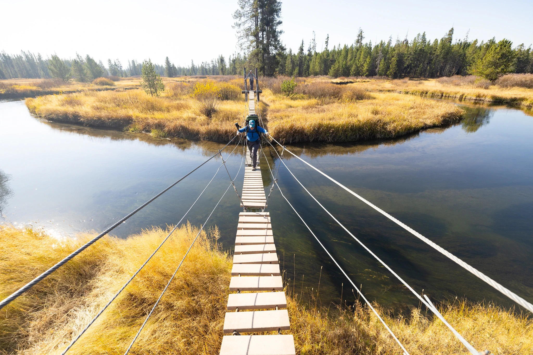 Two people carrying large backpacks who are walking across a wooden suspension bridge that crosses a river.