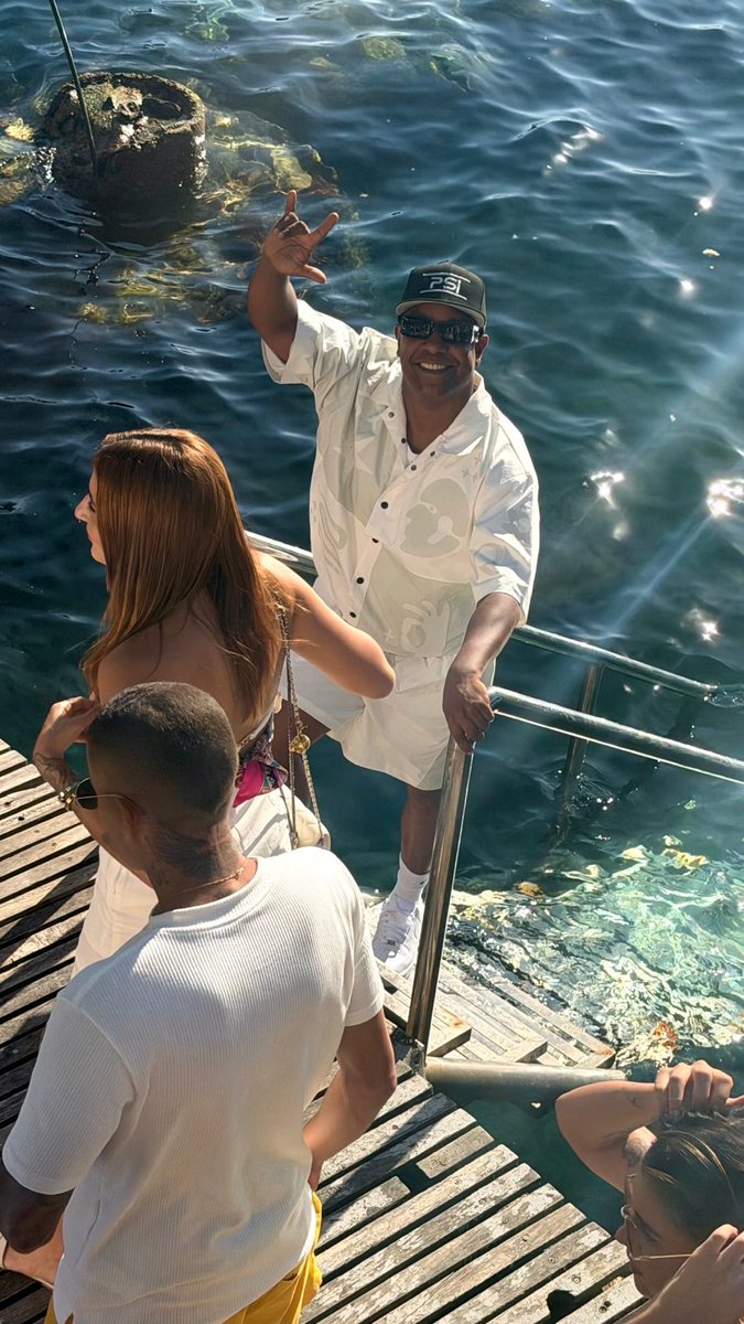 A man in a white patterned shirt and a black 'PSE' cap smiles and waves from a boat dock, with other people nearby and clear water around.
