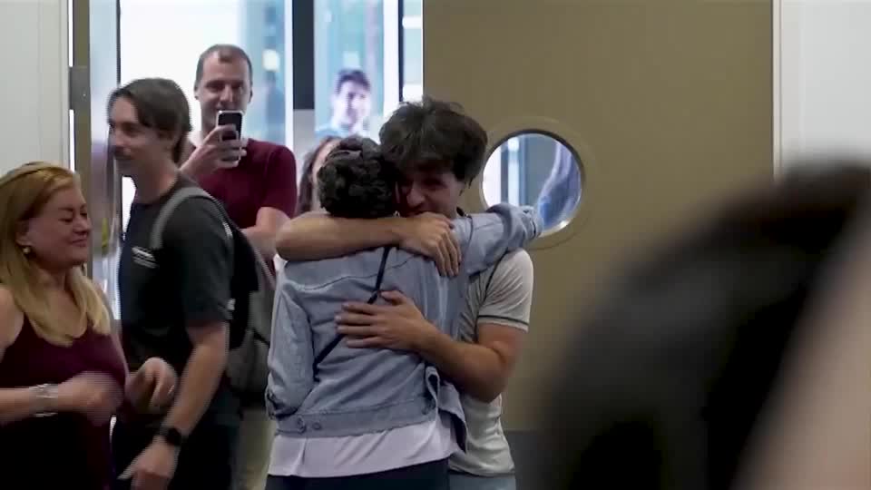 A warm embrace takes place between two people in an indoor setting, with onlookers watching and a man in the background taking a photo with his phone, likely celebrating the Nobel Prize recognition mentioned in the caption. The image does not contain any visible text, numbers, tickers, or names related to the neuroscience achievement.