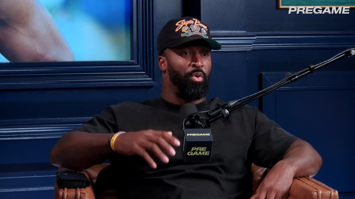 Former NFL player LeBron James speaks into a 'PREGAME' branded microphone while wearing a 'Super Bowl' hat in a podcast studio with dark blue paneled walls.