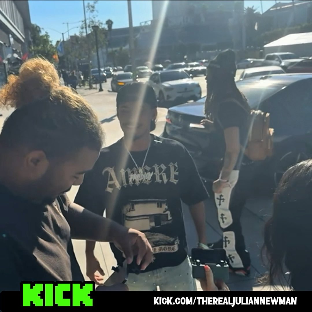 A young man wearing a black "Amore" t-shirt and chains stands on a sunny street at what appears to be ComplexCon, appearing tense while interacting with someone filming him, alongside a woman holding an "FT" bag. The "KICK.COM/THEREALJULIANNEWMAN" banner at the bottom ties to the post about Julian Newman allegedly crashing out on everyone, including his sister's cameraman, as the man in the "Amore" shirt seems to be in a heated moment with a camera operator.