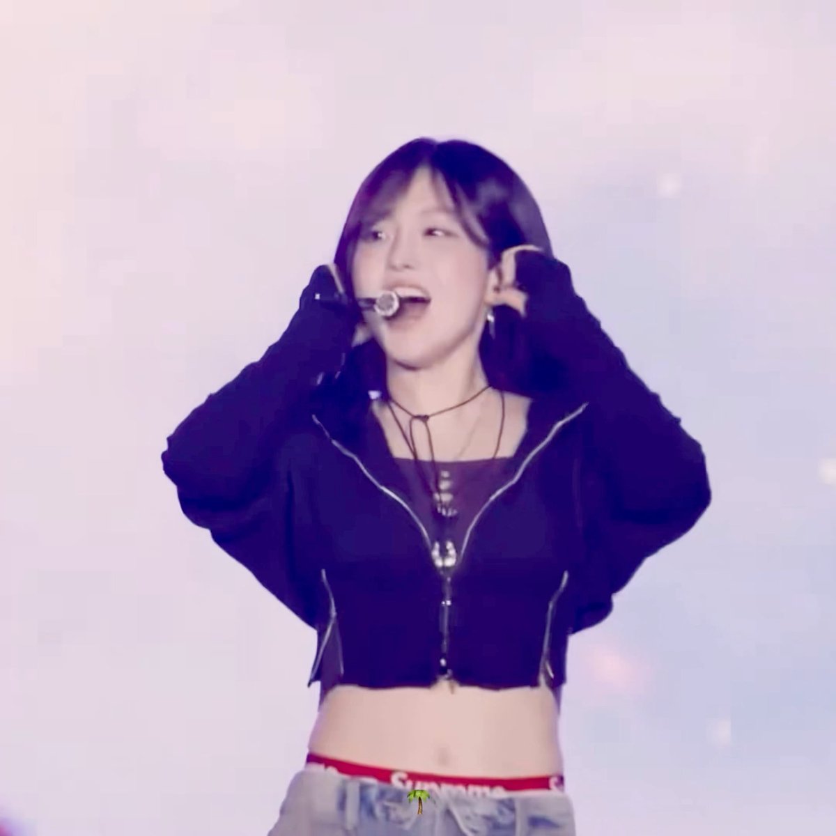 A performer wearing a black zip-up jacket and red 'Supreme' underwear waistband holds both hands up to her head, with a headset microphone on her face against a pinkish background.