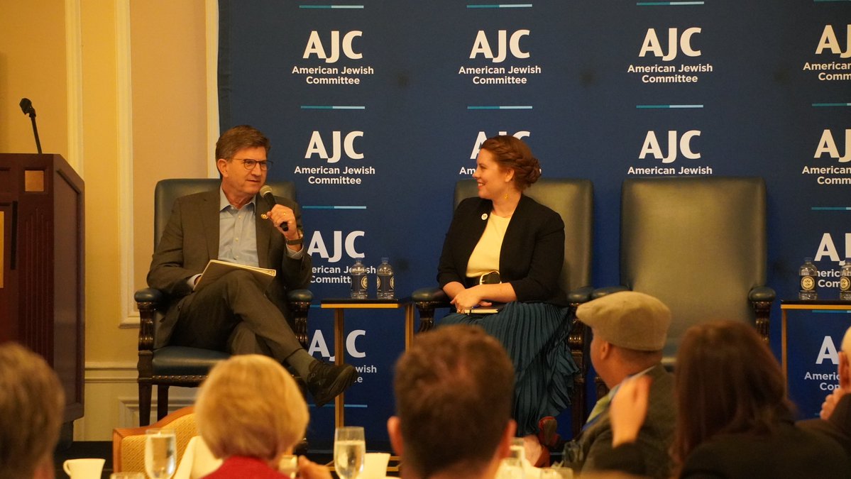 A man and a woman sit in chairs on a stage during a discussion, with an audience in the foreground. A branded backdrop is behind them, and a lectern stands at the left. Text appears on the backdrop.