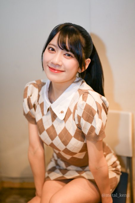 A young woman with a ponytail and bangs wearing a brown and white checkered dress, smiling at the camera, with '@metal_kenji' watermark visible.