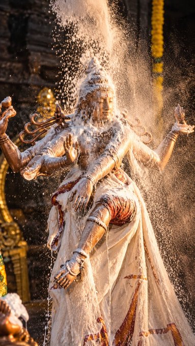 A bronze statue of a multi-armed Hindu deity (Nataraja) being drenched with water or milk, with streams cascading over the figure against a golden temple backdrop.