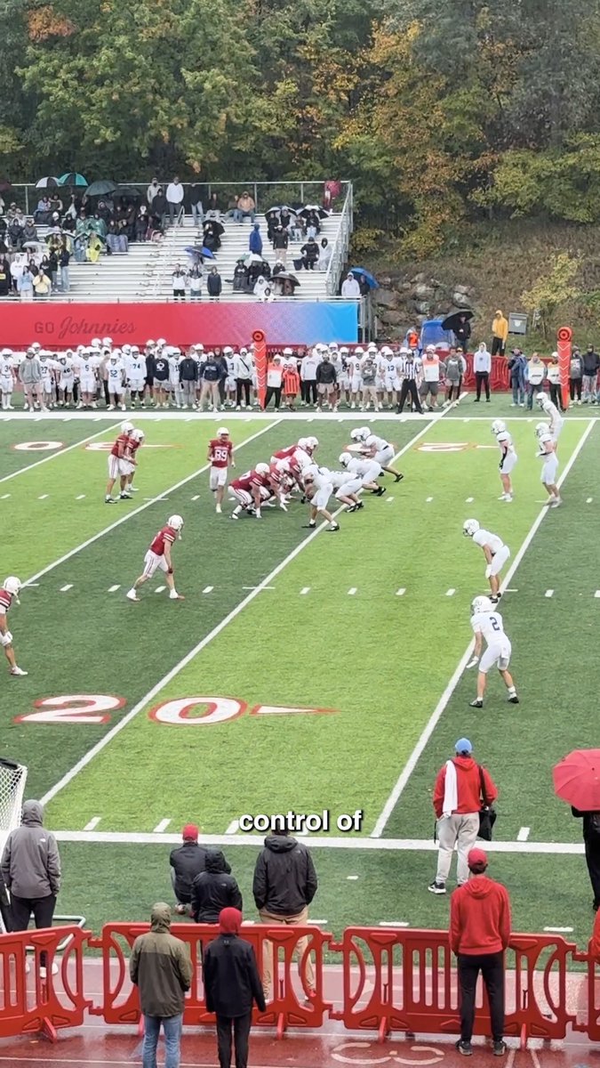 A high school football game in progress on a rainy day with fans in the stands under umbrellas; text overlay reads 'control of', with a banner in the background saying 'Go Johnnies'.