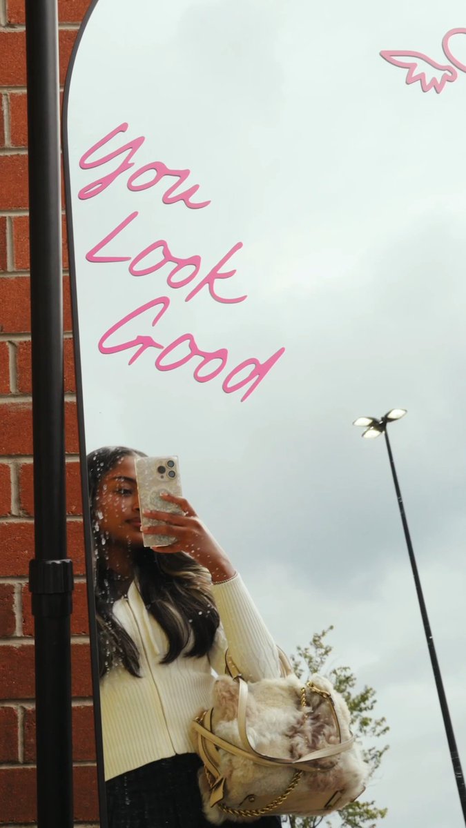 A reflection in an outdoor mirror with pink text reading 'You Look Good' shows a woman in a yellow cardigan taking a mirror selfie with a phone, accompanied by a fluffy handbag.